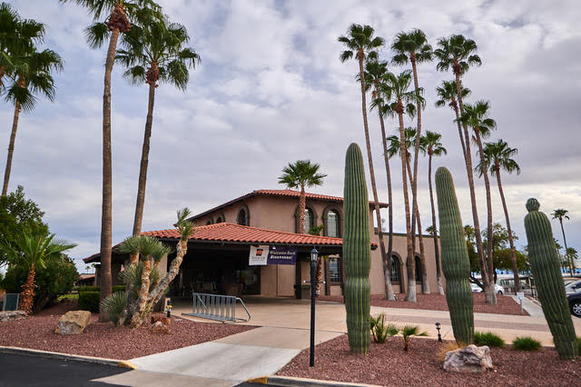 Single-story building with a red tile roof surrounded by tall palm trees and cacti under a cloudy sky; bike rack and sign visible near the entrance.