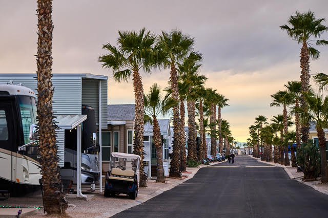 A paved road runs through an RV park lined with palm trees, with motorhomes, small buildings, and golf carts parked along both sides.