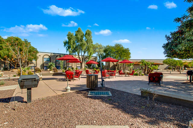 Outdoor patio area with red umbrellas, seating, a barbecue grill, trash can, and surrounding desert landscaping under a clear blue sky.