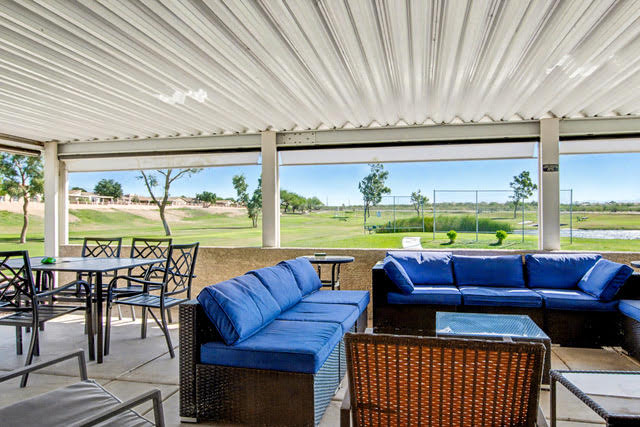 Covered outdoor patio with blue cushioned sofas and black dining tables, overlooking a grassy golf course and tennis courts on a sunny day.