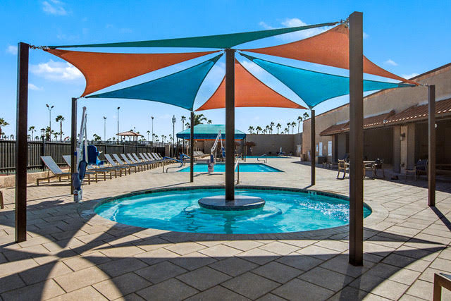 A small round pool with overhead shade sails, surrounded by lounge chairs; a larger rectangular pool is visible in the background on a sunny day.