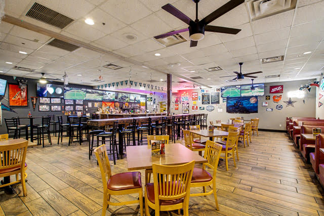 Casual sports bar interior with wooden tables and chairs, red booth seating, wall decorations, ceiling fans, and a long bar counter.