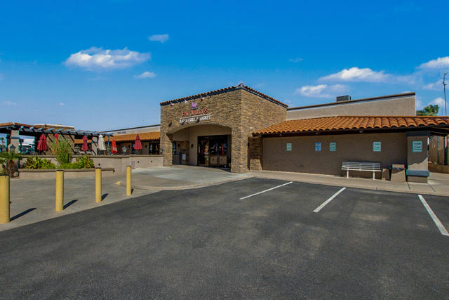 Single-story restaurant with stone entrance, outdoor seating area with umbrellas, empty parking spaces, and clear blue sky.