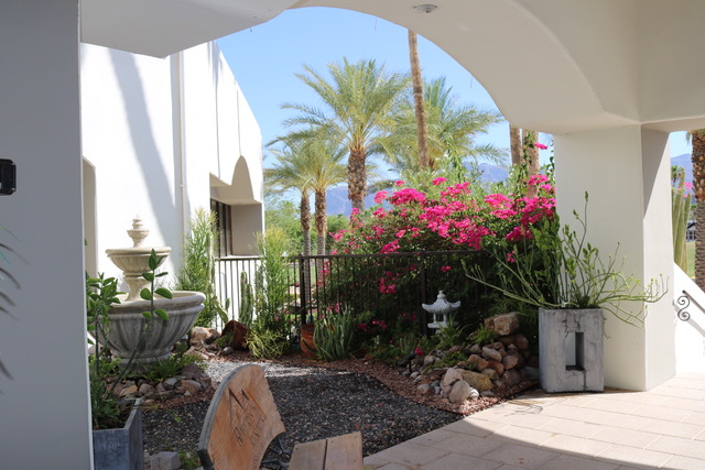 A shaded patio area overlooks a small garden with a fountain, flowering pink bougainvillea, palm trees, and views of mountains in the background.