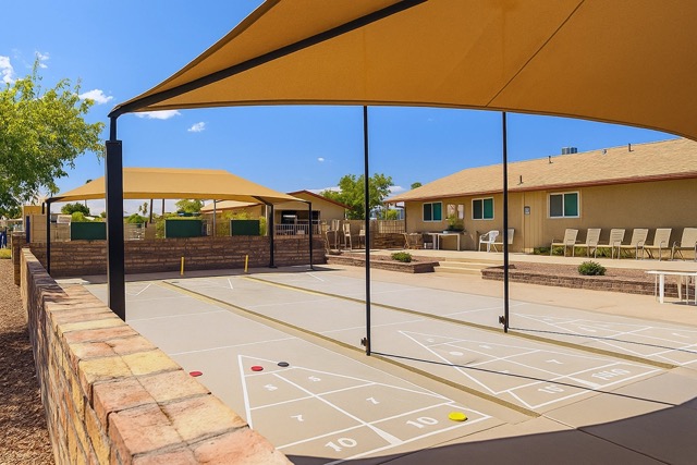 Outdoor shuffleboard courts with shaded canopies, situated next to a beige building and surrounded by desert landscaping under a clear blue sky.