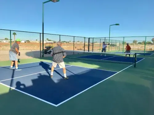 Four people play doubles pickleball on an outdoor court under a clear blue sky, surrounded by a chain-link fence.