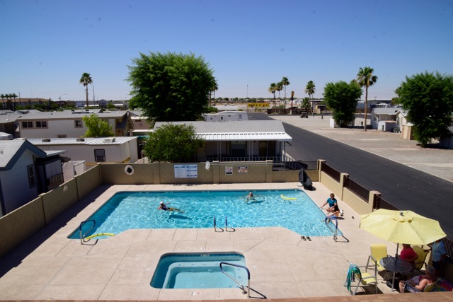 A rectangular outdoor pool with a few swimmers, a hot tub in front, and people sitting under a yellow umbrella in a residential area with palm trees and clear skies.