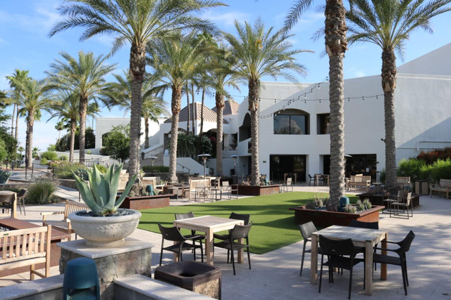 Outdoor patio area with tables and chairs, surrounded by palm trees and modern white buildings, under a blue sky.