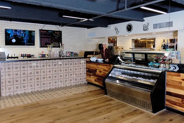 Modern café interior with a patterned tile counter, wood accents, glass display case, menu boards, and a wall-mounted TV. The floor combines wood and decorative tiles.