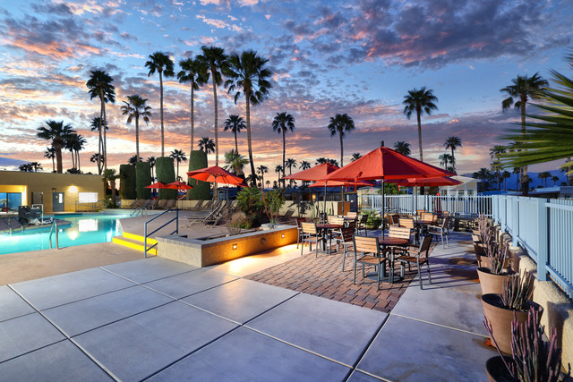 Outdoor dining area with wooden tables and red umbrellas beside a swimming pool, surrounded by palm trees at sunset.