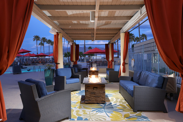 Covered outdoor lounge area with cushioned wicker seating, fire pit table, patterned rug, and red curtains; poolside setting with palm trees and red umbrellas in the background.