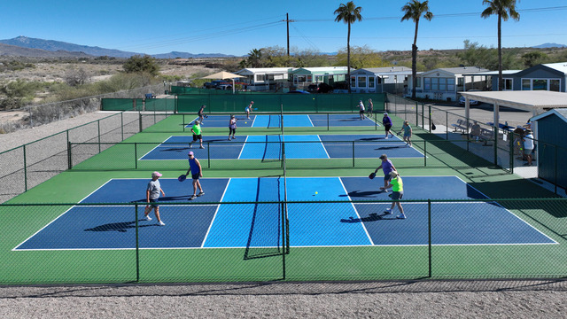 A group of people play pickleball on four outdoor courts surrounded by fencing, with mobile homes and palm trees visible in the background.