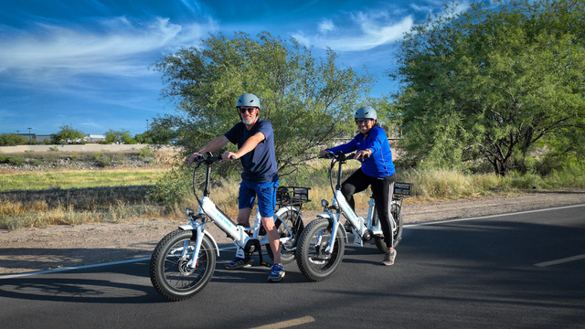 Two people wearing helmets ride electric bikes on a paved path surrounded by trees and dry grass under a blue sky with wispy clouds.