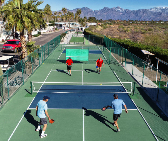 Four men play pickleball on adjacent outdoor courts surrounded by fencing, palm trees, and a mountain range in the background.