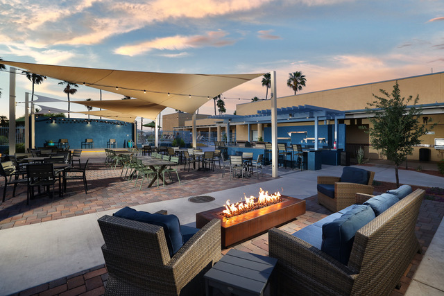 Outdoor patio with cushioned seating around a rectangular fire pit, dining tables, shade sails, and palm trees at sunset.