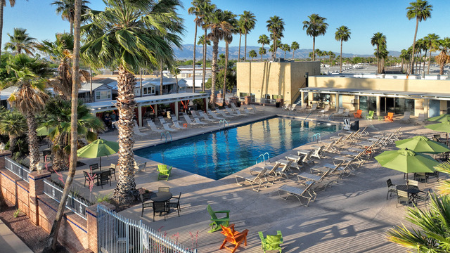 Outdoor swimming pool surrounded by lounge chairs, palm trees, and tables with green umbrellas, next to a building with mountains and blue sky in the background.