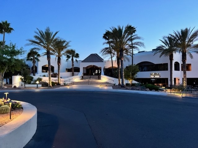A large, white, modern building with arched windows and palm trees at the entrance, photographed at dusk with exterior lights illuminating the walkway.
