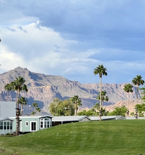 Green lawn with palm trees and small white homes in the foreground, mountains and a partly cloudy sky in the background.