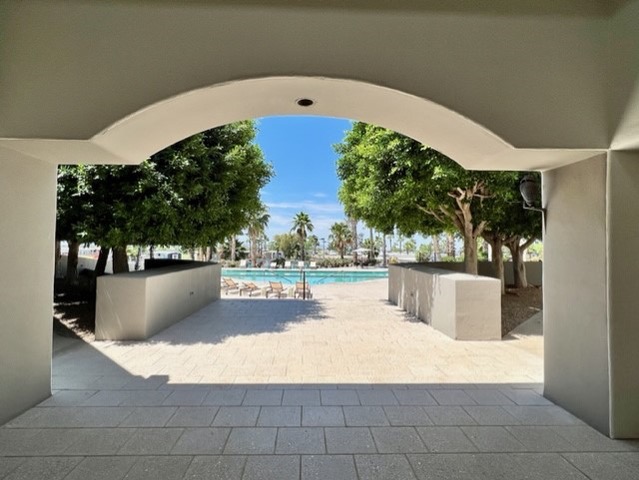 View through an arched walkway showing a patio area with lounge chairs, trees, and a swimming pool under a clear blue sky.
