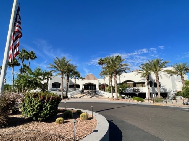 A white, modern building with large arched windows is surrounded by palm trees and desert landscaping under a clear blue sky. An American flag stands in the left foreground.