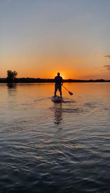 A person paddles on a stand-up paddleboard across calm water at sunset, with the sun near the horizon and trees in the distance.