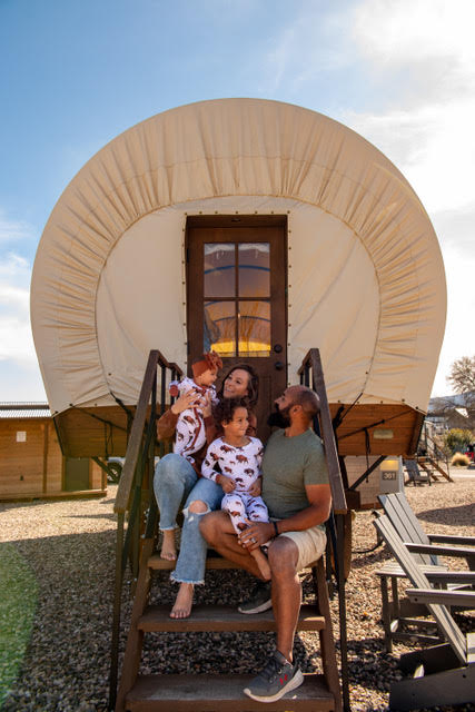 A family of four sits on the steps of a covered wagon cabin, smiling and wearing matching pajamas, with a clear sky in the background.