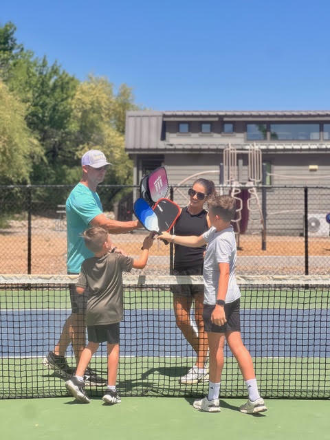 Four people on a tennis court, two adults and two children, stand near the net touching their rackets together in the center.
