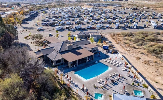 Aerial view of a large RV park with numerous parked RVs, a central building, an outdoor swimming pool, lounge chairs, and two hot tubs, surrounded by dry vegetation.
