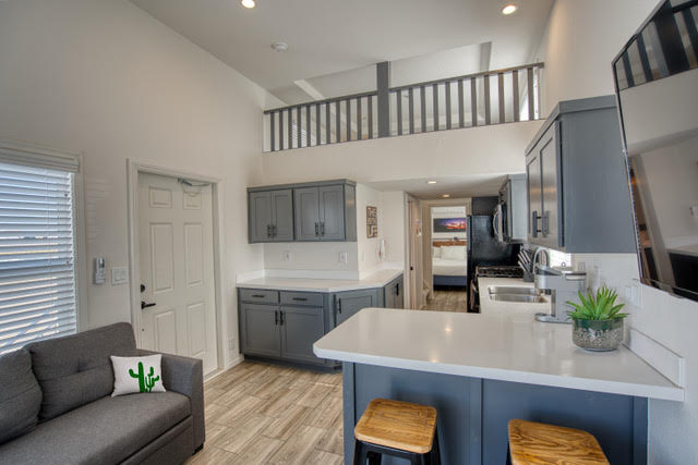 Modern kitchen and living area with gray cabinets, white countertops, wood stools, loft railing above, and a partially visible bedroom in the background.