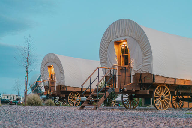 Two covered wagons with lit interiors are parked side by side on gravel, each with stairs leading up to the entrance, during early evening.