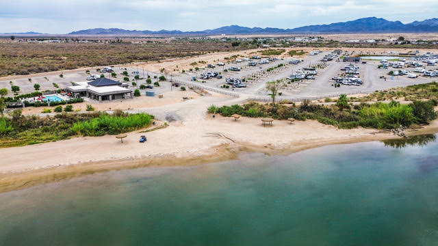 Aerial view of a sandy beach by a calm river, with a parking lot, small building, and RVs in a desert landscape under a cloudy sky.