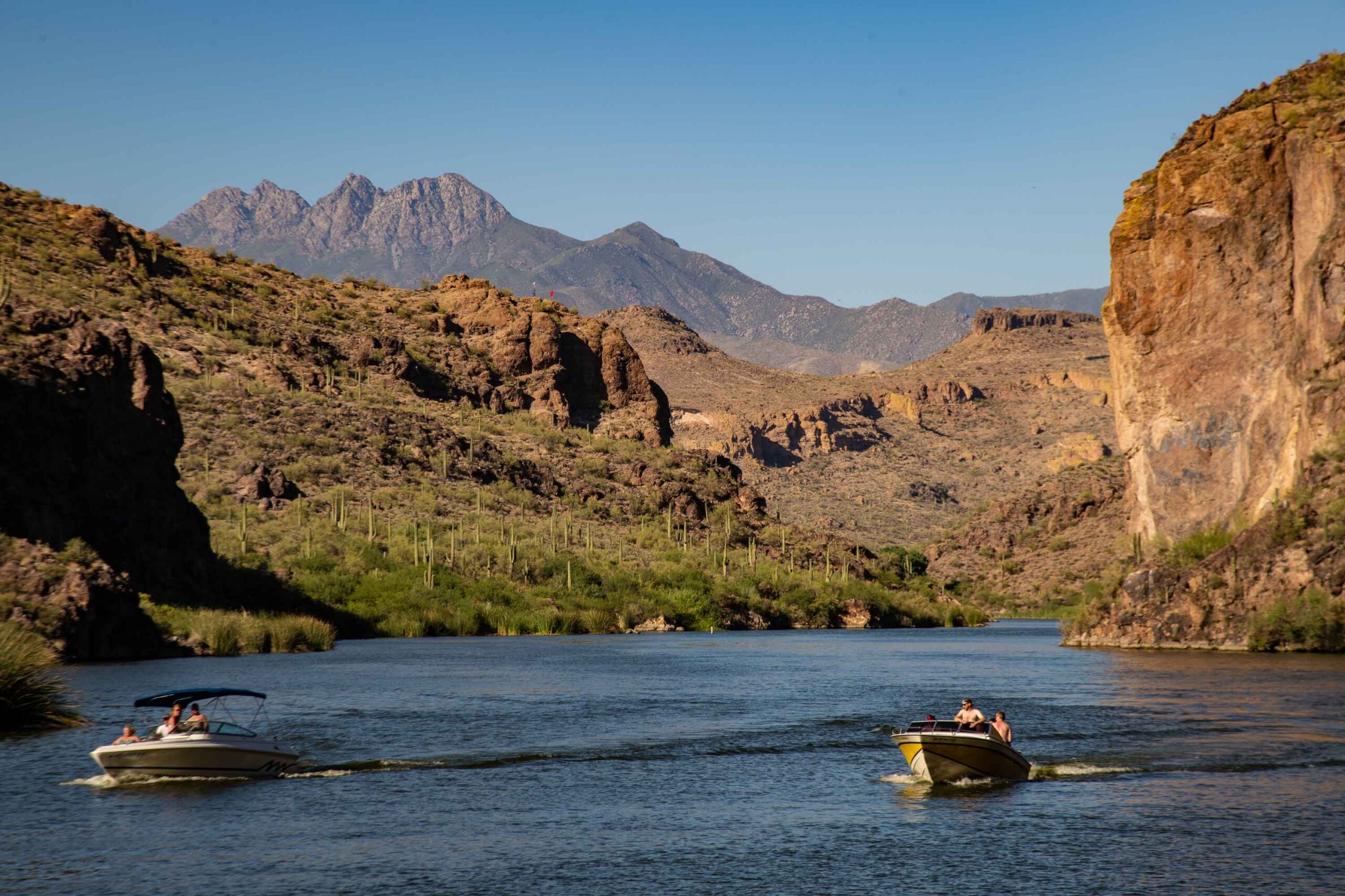 Two motorboats travel along a river surrounded by rocky cliffs and desert vegetation, with mountains visible in the background under a clear blue sky.