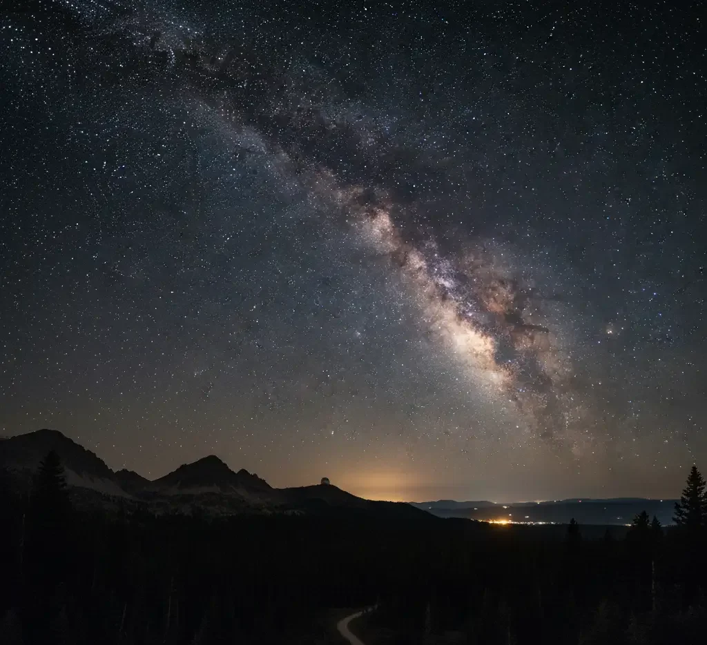 A wide view of the Milky Way galaxy stretches across a star-filled night sky above silhouetted mountains and distant city lights.